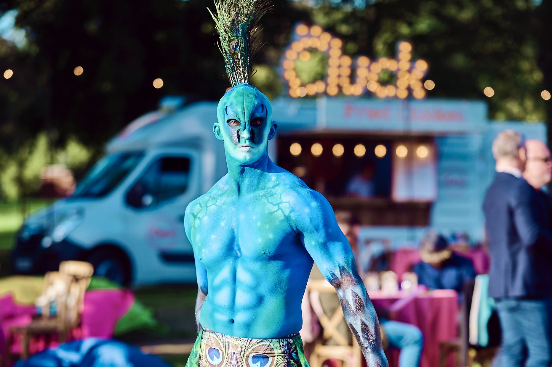 Peacock A toned male circus performer body painted with prosthetics and feathered headdress as a peacock at a summer garden party on a private estate